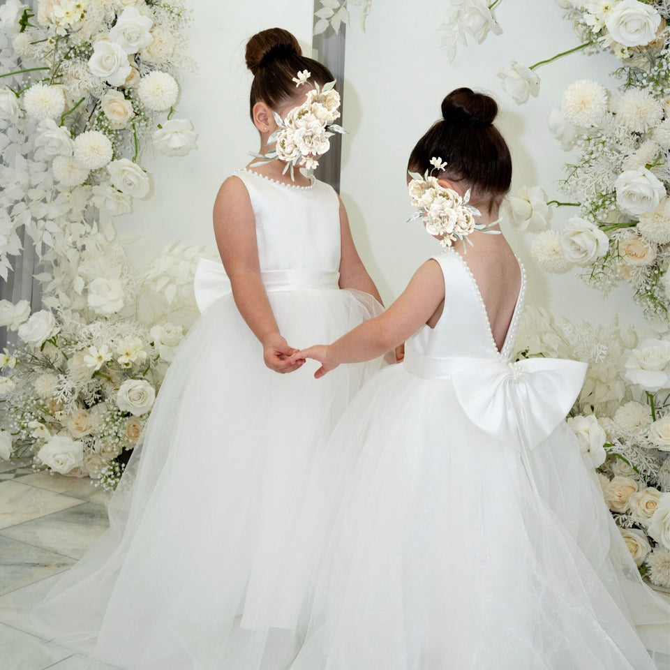 Two flower girls in their floor length dresses with big bows and open backs are standing and holding hands in front of decorated flower background